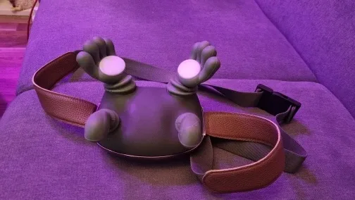 Das Massagegerät von oben auf einem Sofa in violettem Licht, die beiden Silikonhände mit ihren hellen Wärmepads nach oben, Lederriemen und Schultergurt daneben ausgebreitet
