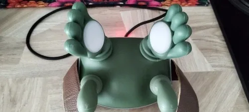 Das grüne Massagegerät von vorne auf einem Holzboden, beide Silikonhände geöffnet, die weißen Wärmepads in den Handflächen sichtbar, das Ladekabel dahinter