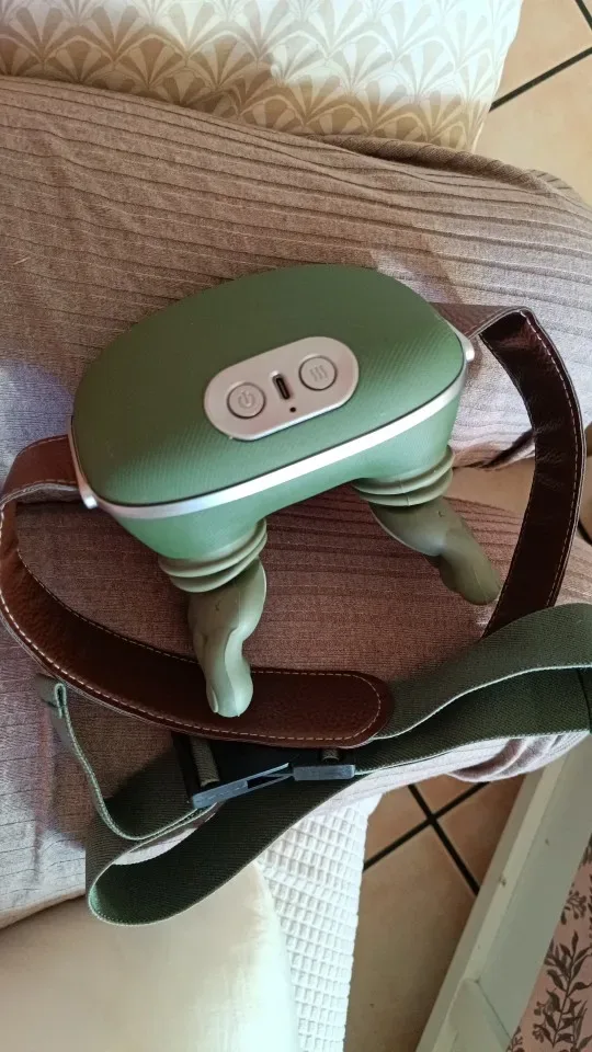 Das Massagegerät auf einem beigen Sofa, der braune Lederriemen um das Gehäuse gelegt, der Chromring beleuchtet